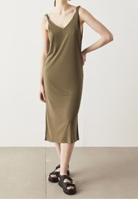Robe vert olive, à mi-mollet, avec un décolleté en V et des bretelles nouées ; portée avec des sandales noires chunky ornées d'accents décoratifs.