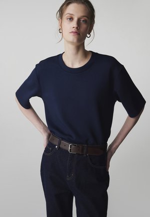 Jonge vrouw die een donkerblauw oversized T-shirt draagt, ingestopt in een donkere spijkerbroek met een bruine riem, staand met handen in de zij, neutrale achtergrond.