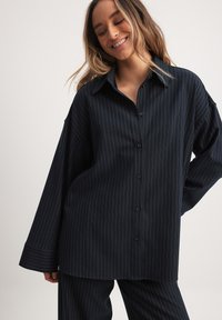 Camicetta a maniche lunghe di colore blu navy con strisce verticali bianche. Presenta un colletto classico e una vestibilità comoda con polsini larghi. Materiale morbido.