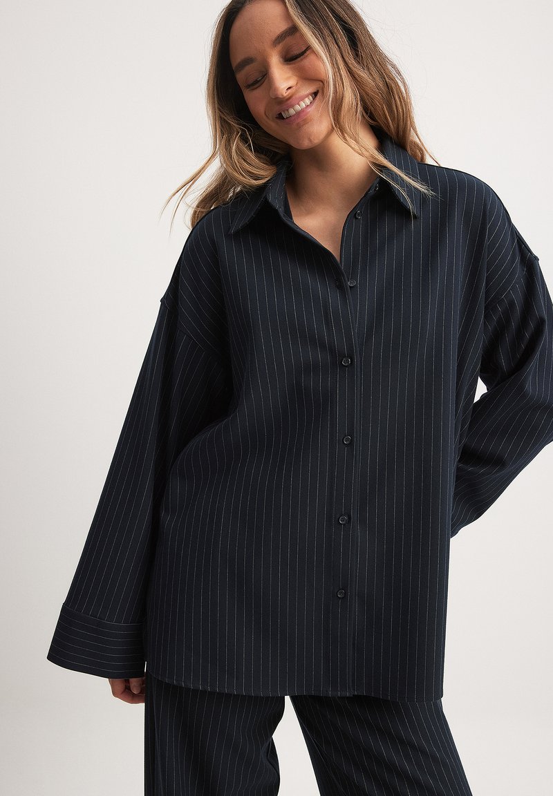 Melnbalti svītrainā pogājama krekliņa ar apkakli, pārāk liels piegriezums un garām piedurknēm, izgatavots no gludas auduma.