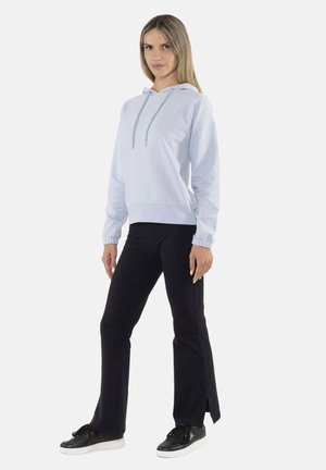 Femme debout les mains détendues, portant un sweat à capuche bleu clair, un pantalon noir avec des fentes latérales et des baskets noires à semelles blanches.