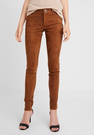 Pantalon en daim marron coupe ajustée porté avec un haut beige et des escarpins noirs à bout ouvert sur un fond clair.