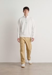 Hvit zip-hals genser, lysebeige bukser og hvite sko. Modellen står mot en enkel bakgrunn og viser frem antrekkets enkle design.