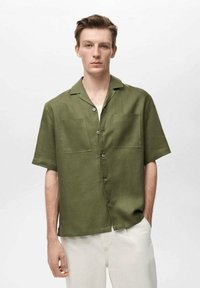 Short-sleeved olive green shirt made of linen, featuring two chest pockets and a button-up design, paired with light-colored pants.