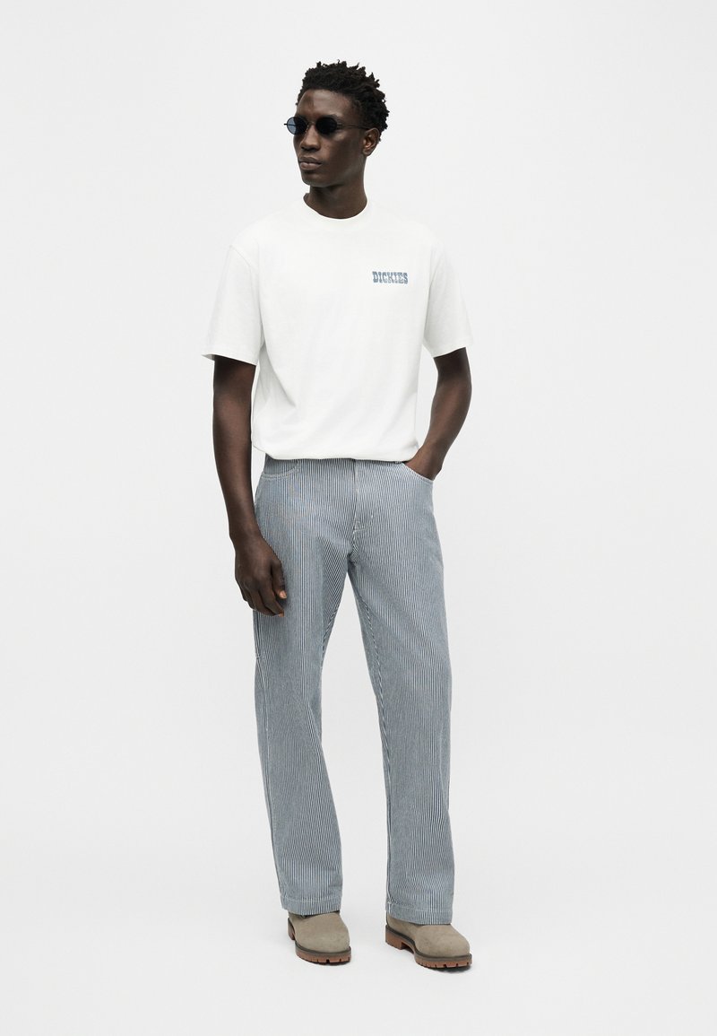 Man wearing sunglasses, white short-sleeve Dickies t-shirt, gray striped pants, and beige boots, standing with one hand in pocket.