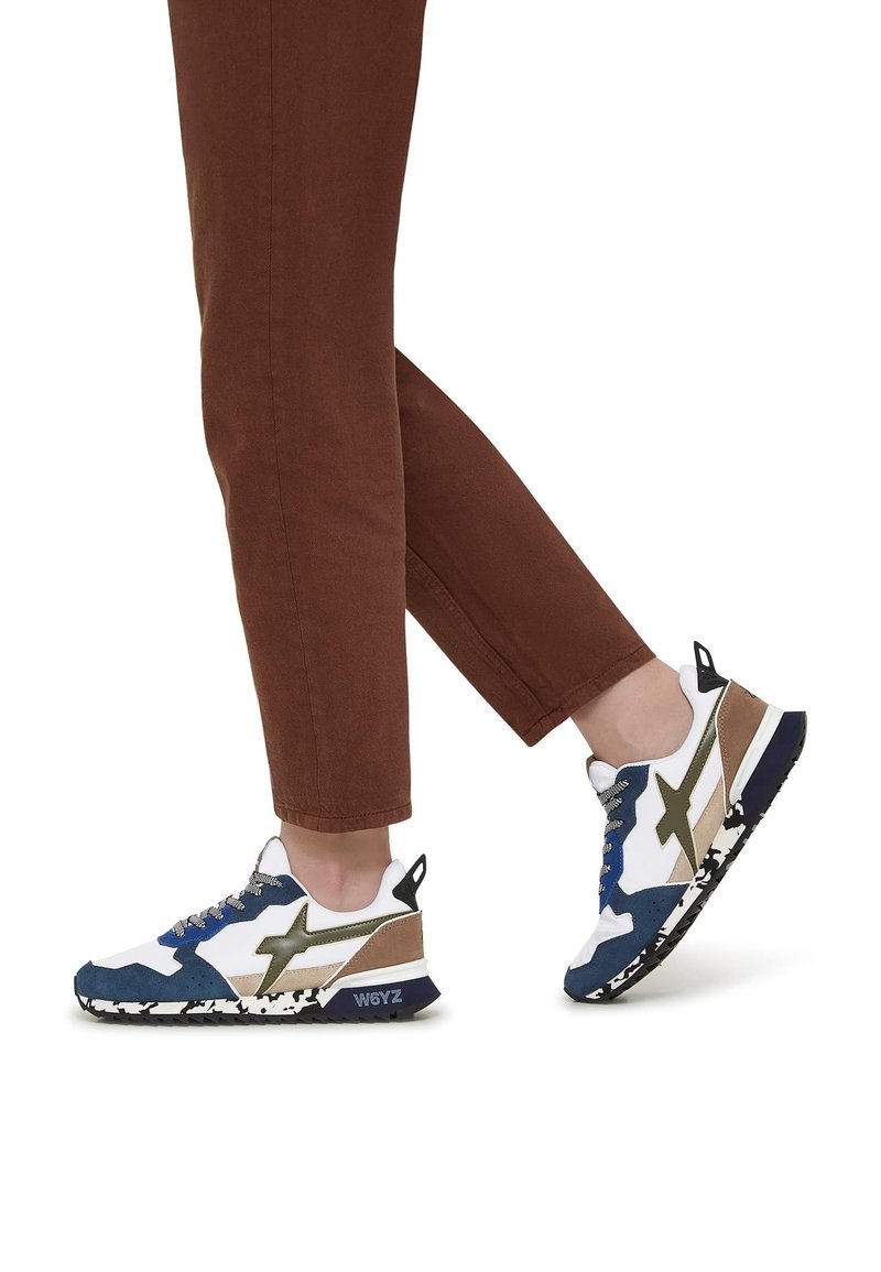Personne portant un pantalon marron et des baskets bleues, blanches et beiges avec des semelles à motifs, marchant sur un fond blanc.