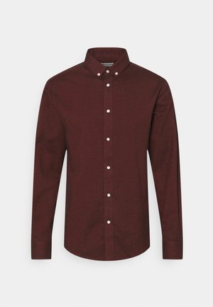 Chemise à manches longues pour homme, boutonnée, de couleur bordeaux foncé avec des boutons blancs et un col pointu, sur un fond uni.