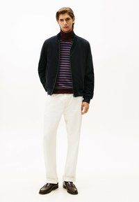 Chaqueta bomber marina con cremallera al frente, llevada sobre un cuello de tortuga burdeos y a rayas, combinada con pantalones blancos y mocasines marrones.