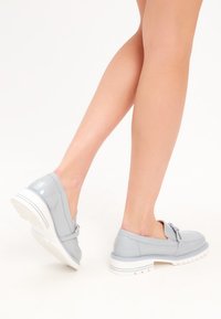 Vaaleanharmaat patentoidut slip-on -kengät, joissa on pyöreä kärki, nauhaaksentti ja teksturoitu valkoinen pohja. Sileä pinta, jossa on hienovaraisia ompeluyksityiskohtia.