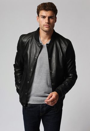 Schwarze Lederbomberjacke mit geripptem Kragen, Druckknöpfen und gesteppten Ärmeln, kombiniert mit einem hellgrauen Pullover und dunkelblauen Jeans.