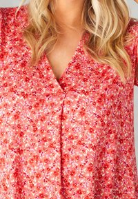 Blusa floral en coral claro con pequeñas flores rojas y rosas. Presenta un escote en V y está confeccionada en una tela suave y ligera.