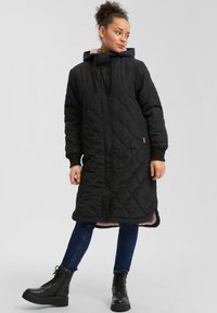 Zwarte gewatteerde lange jas met een capuchon, voorzien van een roze voering, geribbelde manchetten en zijslitsen, gecombineerd met een donkerblauwe spijkerbroek en zwarte veterschoenen.