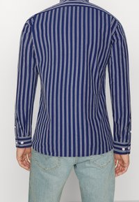 Uomo che indossa una camicia a maniche lunghe blu con strisce verticali bianche e jeans azzurri chiari, in piedi con le spalle rivolte verso la fotocamera.