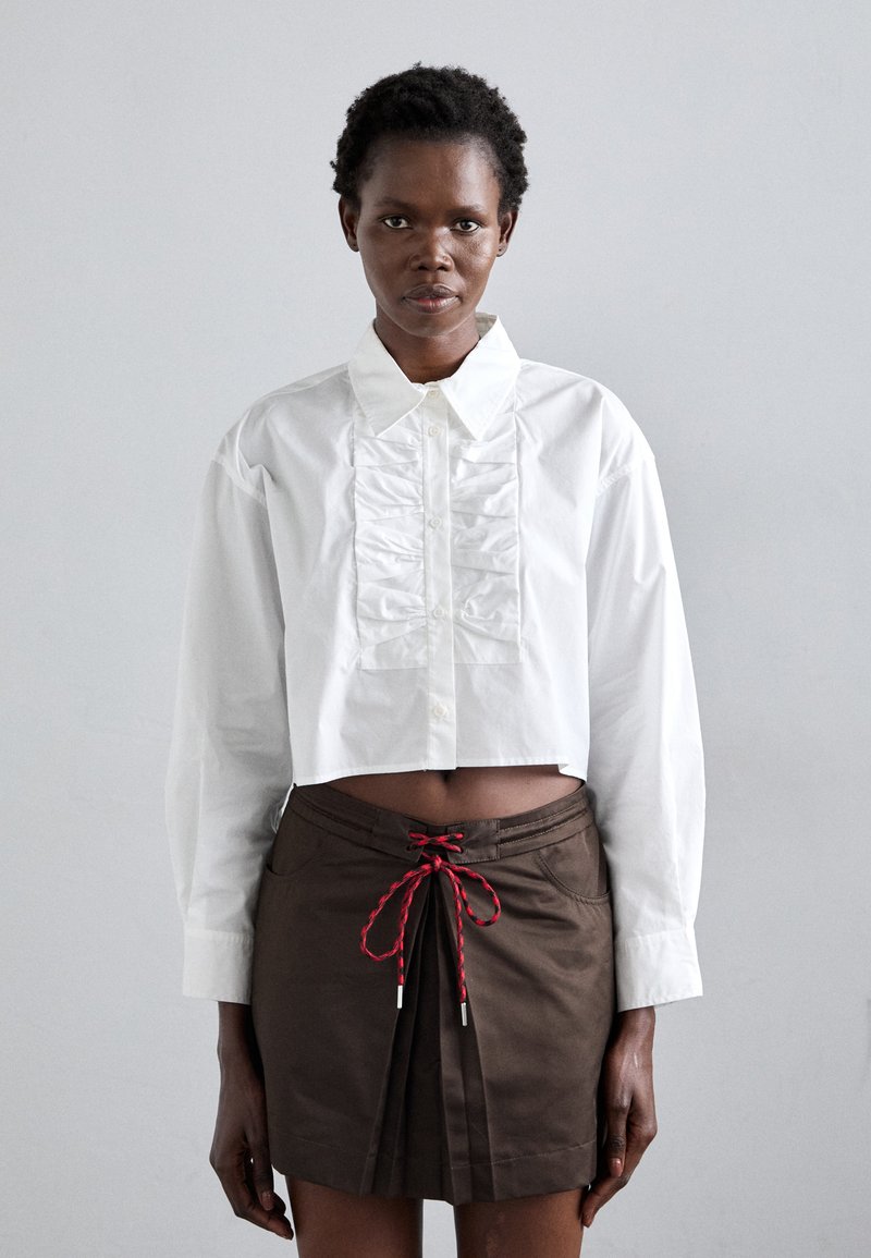 Chemise blanche courte avec un détail froncé à l'avant, associée à une jupe marron à plis et à une fermeture à cordon rouge. Tissu en coton texturé.
