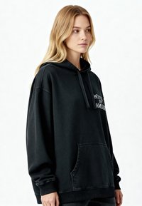 Fekete, túlméretezett kapucnis pulóver elülső zsebbel, bordázott mandzsettákkal, és hímzett szöveggel: "HÔTEL DE AMOUR." Puha anyag, amely texturált megjelenésű.