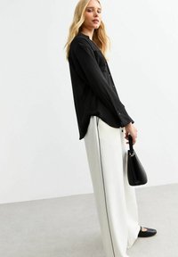 Zwarte overhemd met lange mouwen, gecombineerd met witte wijde broek met een zwarte zijstreep. Accessoires omvatten een kleine zwarte handtas.
