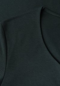Tmavě zelená, měkká bavlněná tkanina zblízka představuje hladký povrch a výstřih ve tvaru lodi. Minimalistický design bez viditelného kovového spojení nebo vzorů.