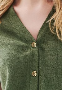 Grüner gestrickter Cardigan mit V-Ausschnitt, strukturiertem Stoff und einem einzelnen goldenen Knopf am Kragen.