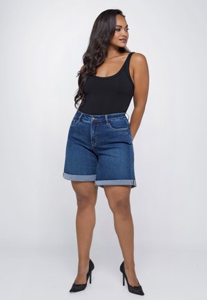 Woman with long hair wearing a black tank top, rolled-up blue denim shorts, and black high heels standing against a plain background.