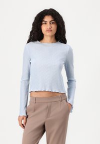 Haut long à manches en bleu clair, à côtes, avec un ourlet court et une finition texturée, associé à un pantalon tailleur marron pour un contraste.