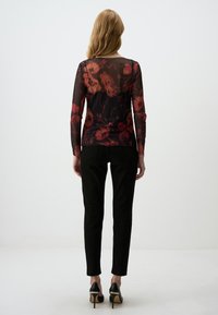 Haut long à manches transparentes noir avec un motif floral rouge, associé à un pantalon slim noir et des chaussures à talons hauts noires. Fond uni.