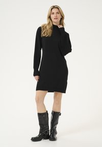 Schwarzes Strickkleid mit langen gerippten Ärmeln, hohem Kragen und figurbetonter Silhouette. Kombiniert mit hohen schwarzen Lederstiefeln mit Schnallen.