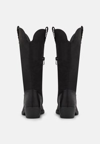 Botas altas negras de ante texturizado, con un borde superior ondulado, correas ajustables y un tacón apilado para mayor estabilidad.