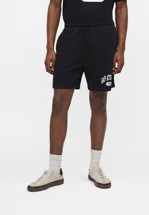 Αρσενικό μοντέλο φοράει μαύρα σορτς με κορδόνι και το κείμενο "GAP ATHL 1969" στο αριστερό πόδι, λευκές κάλτσες και μπεζ αθλητικά, στέκεται μπροστά σε λευκό φόντο.