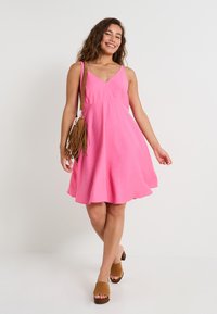 Vestido rosa sin mangas con escote en V y falda acampanada, hecho de tela ligera. Combinado con mules de tacón marrones y un bolso bandolera con flecos.
