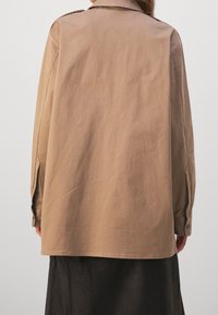 Lysebrun skjortejakke med lange ærmer i glat stof. Har krave, knapmanchetter og en let løs pasform.