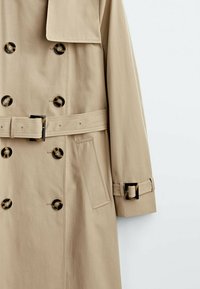 Trench-coat beige avec boutons à double boutonnage en écaille de tortue, taille ceinturée, poche latérale et bride à boucle au niveau du poignet.