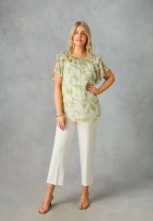 Blusa floreale verde chiaro con maniche a volants, abbinata a pantaloni bianchi a gamba dritta e sandali dorati con cinturini. Tessuto morbido.