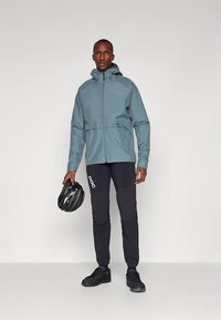 Αδιάβροχο μπουφάν με κουκούλα σε ανοιχτό γαλάζιο, συνδυασμένο με μαύρο παντελόνι και μαύρο κράνος ποδηλάτου. Διαθέτει εμπρόσθιο φερμουάρ και ρυθμιζόμενες μανσέτες.