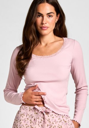 Vrouw met lang bruin haar die een lichtroze top met lange mouwen en kanten rand draagt, en roze pyjamabroek met bloemenpatroon en een gestrikte strik.