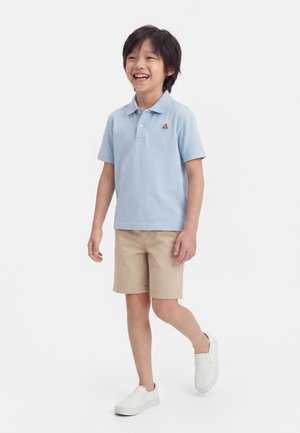 Băiat zâmbitor care merge, purtând un tricou polo albastru deschis cu un mic logo urs, pantaloni scurți bej și pantofi slip-on albi pe fundal alb.