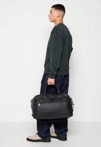 Černá taška typu duffel z odolného materiálu, obdélníkového tvaru, s texturovaným povrchem, horními rukojetěmi a kapsou na zip na přední straně.