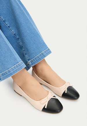 Füße in cremefarbenen Ballerinas mit schwarzen Zehenkappen und kleinen Schleifen, kombiniert mit blauen Jeans.