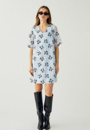 Kobieta w jasnoniebieskiej sukience w kwiaty, czarnych okularach przeciwsłonecznych i czarnych, teksturowanych botkach za kolano, stojąca na tle gładkiego, jasnego tła.