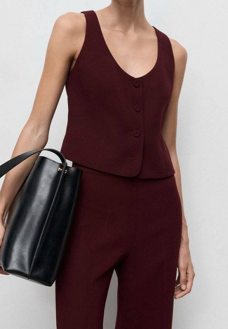 Femme portant un haut bordeaux sans manches boutonné et un pantalon assorti, tenant un sac à main en cuir noir avec une seule bandoulière.