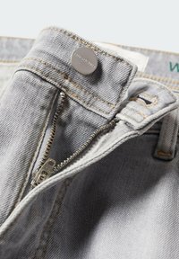 Jean en denim gris clair avec fermeture éclair métallique partiellement ouverte et bouton métallique gravé du nom de la marque "MNGDENIM".