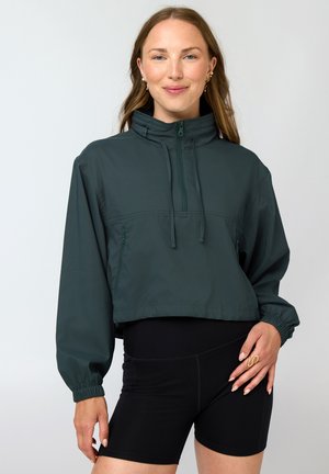 Naeratav naine, kes kannab tumerohelist lühendatud lukuga džemprit ja musti kõrge vöökohaga rattapükse, poseerides ühevärvilise tausta ees.