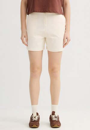 Persona que lleva pantalones cortos color crema, calcetines blancos, zapatillas marrones y una camiseta corta marrón parado frente a un fondo blanco liso.