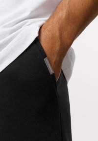 Černé bavlněné tepláky s boční kapsou a stříbrnou cedulkou. Lem je hladký a látka se zdá být mírně texturovaná.