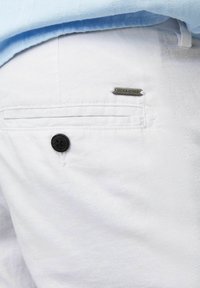 Närbild på en vit byxficka med svart knapp och liten metallisk Jack & Jones-logotyp under fållen på en ljusblå skjorta.