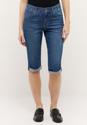 Mujer con pantalones cortos vaqueros de mezclilla azul oscuro hasta la rodilla, con los bajos remangados, y mocasines negros, de pie contra un fondo liso claro.