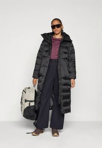 Must pikk talvebinsooniga kapuutsiga mantel, koos burundivarjundse toppi ja tumedate laiade pükstega. Kandmas heledat halli seljakotti ja tumedaid päikeseprille.