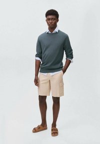 Men's outfit: dark green sweater, light blue collared shirt, beige shorts with side pockets, brown sandals, standing against a plain backdrop.