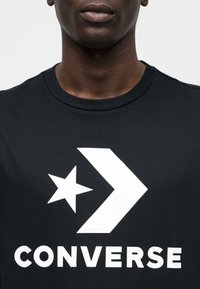 Μαύρο βαμβακερό t-shirt με μεγάλο λευκό λογότυπο Converse που περιλαμβάνει σχεδιασμό με αστέρι και βέλος, στρογγυλή λαιμόκοψη και κοντά μανίκια.