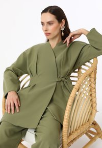 Olive green wrap-style jacket made from smooth fabric, featuring a belted waist and long sleeves. Worn with coordinating pants, seated on a rattan chair.