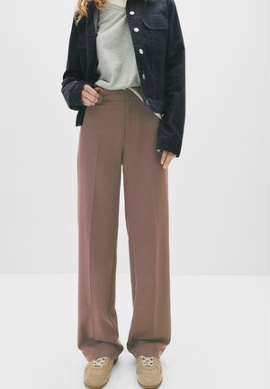 Persoană îmbrăcată cu pantaloni largi maronii, teniși bej, bluză gri și jachetă din denim închis, stând în fața unui fundal alb simplu.
